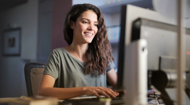 A woman with long brown hair sits at a desk, smiling while using a desktop computer in her home office, researching multiple works registration for copyright protection.