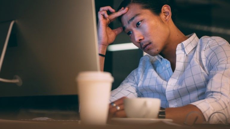A person wearing a white plaid shirt sits at a desk, looking intently at a computer screen—perhaps reviewing strategic filing options or considering single vs. multiple works registration—with a coffee cup in the foreground.