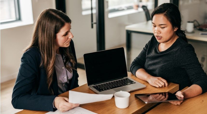 Two women sit at a table in an office, one holding papers and the other a tablet, as they discuss copyright applications, with a laptop and coffee cup between them.